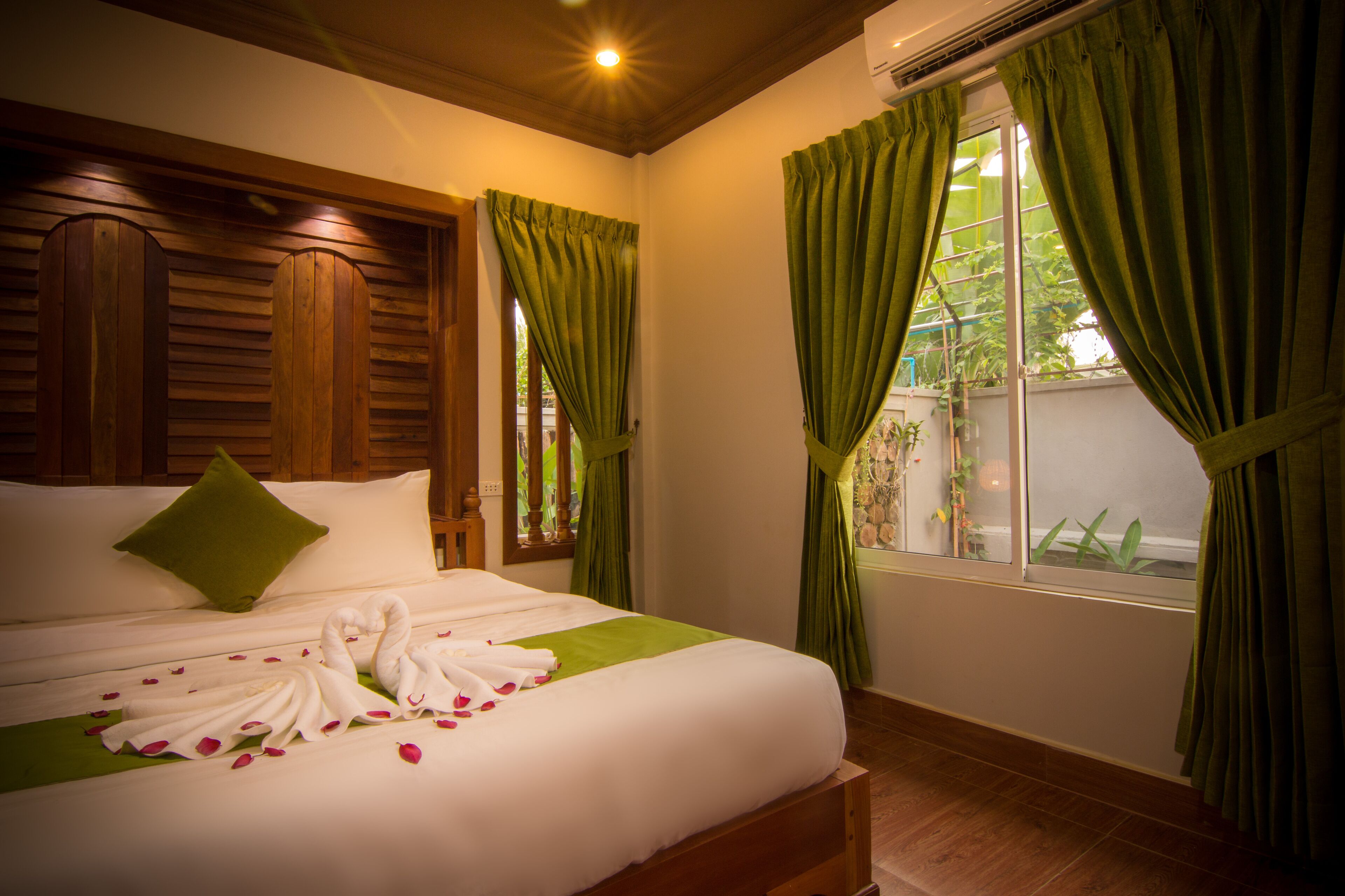 A neatly made bed with white linens and a green bedspread, a green pillow, and a swan-shaped towel arrangement with rose petals.