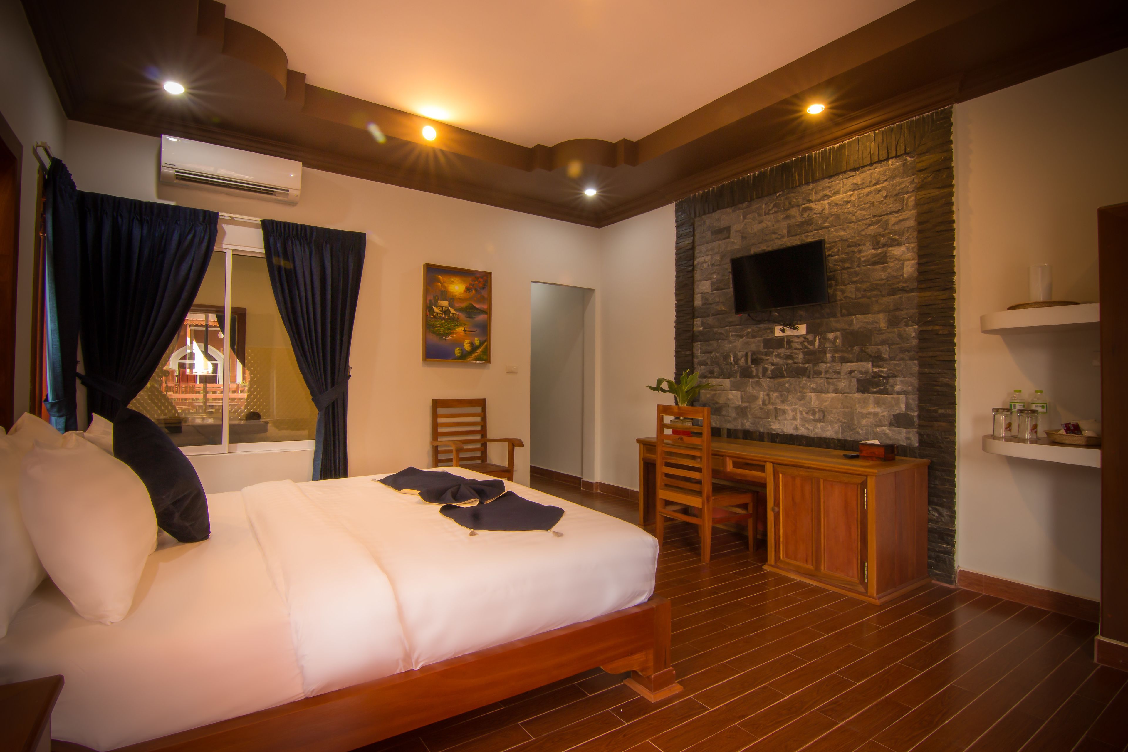 A hotel room with a large bed, wooden furniture, a stone wall, and a flat-screen TV.