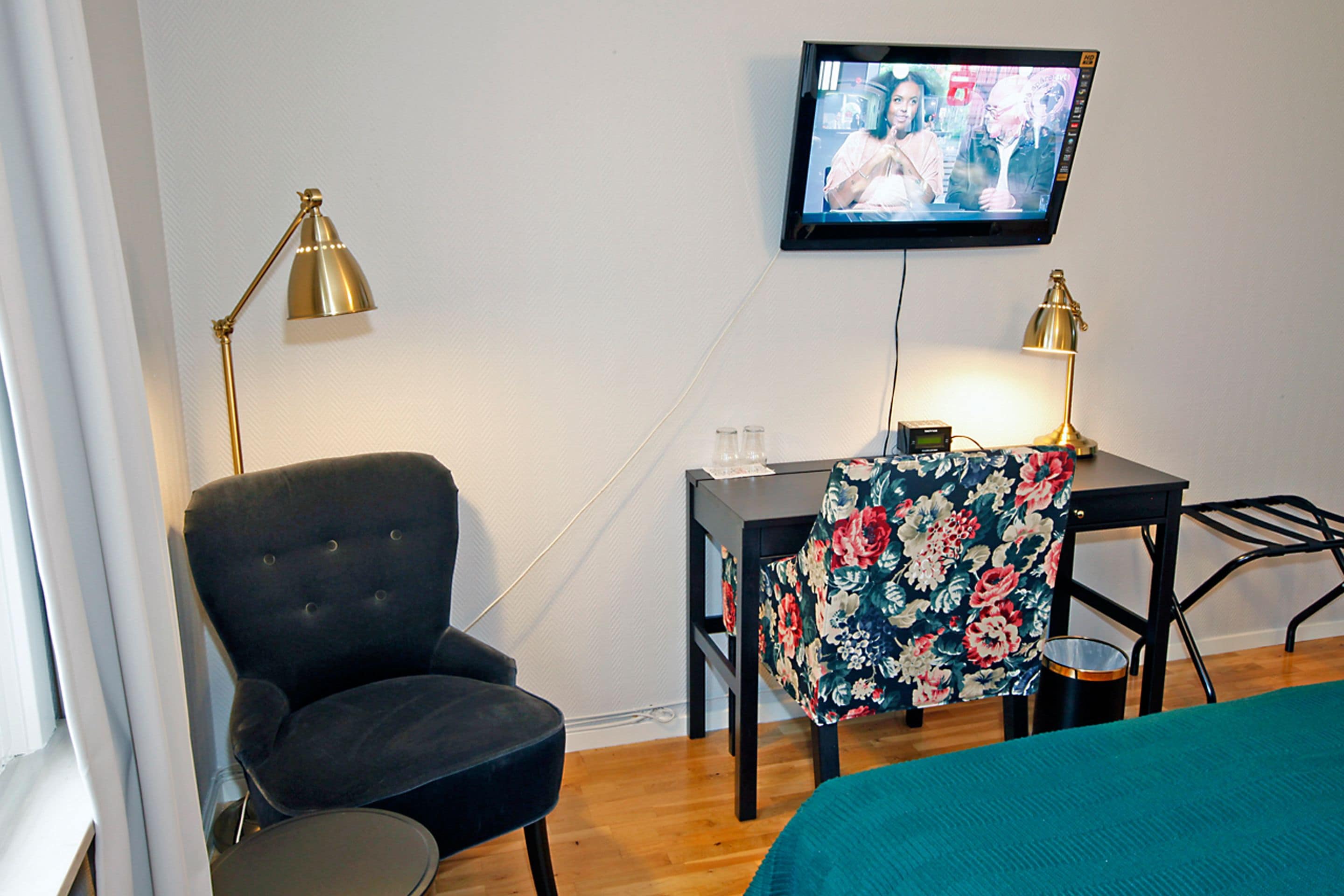 Een hotelkamer met een donkerblauwe fauteuil, een kleine tafel met een bloemig tafelkleed, een aan de muur bevestigde televisie en twee vloerlampen.
