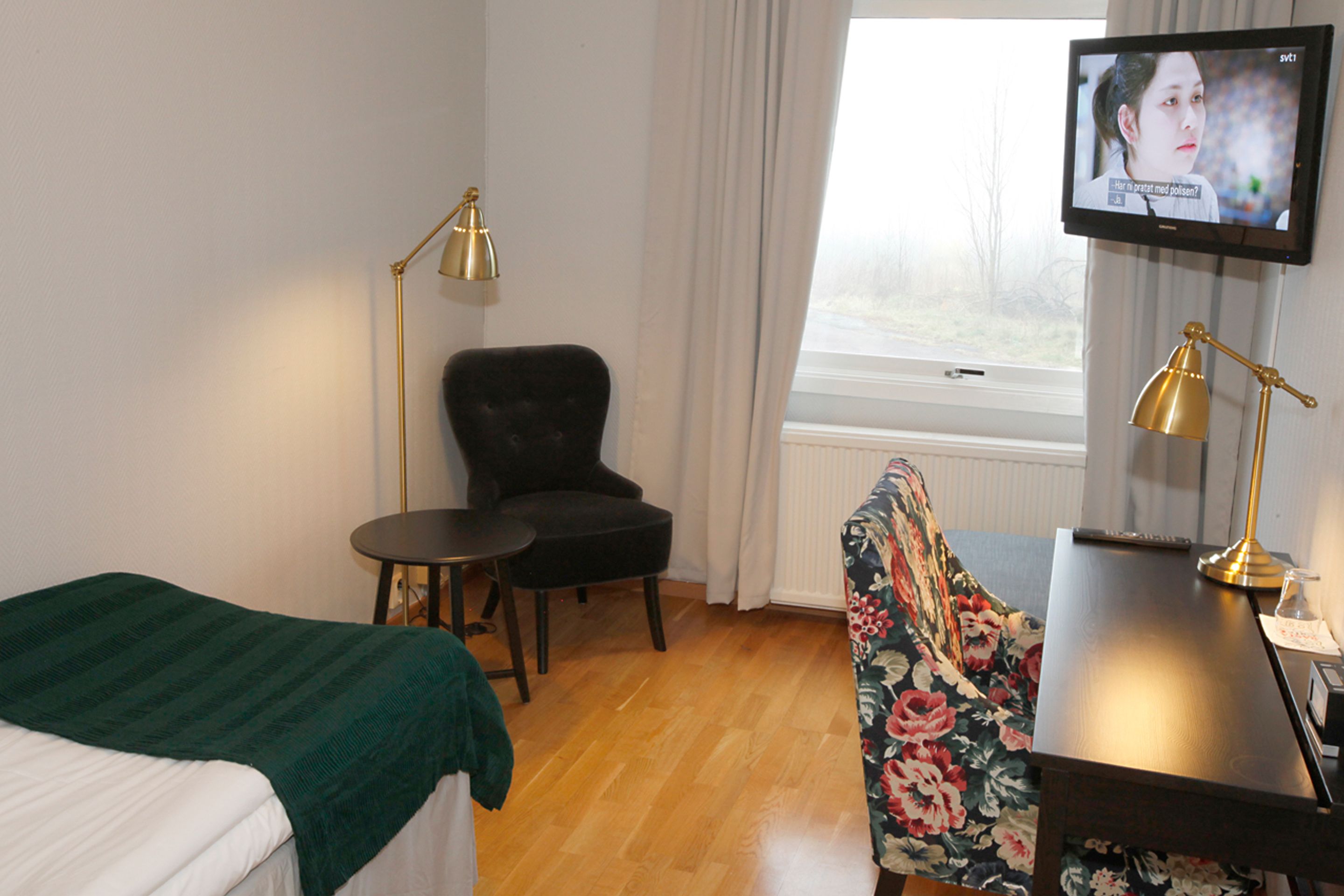 A hotel room with a bed, a chair, a small table, a desk, a TV, and a window with curtains.