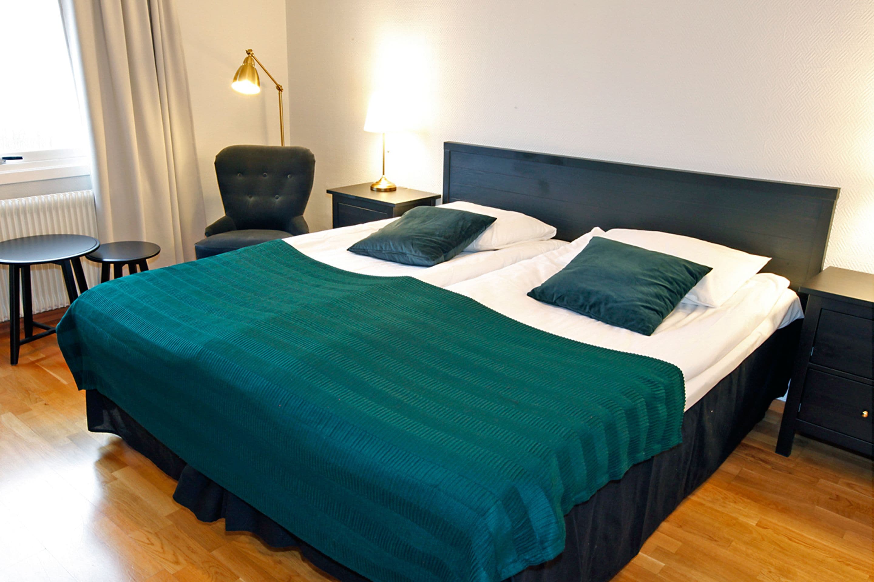 Letto rifatto con copriletto verde e due cuscini, un tavolino, una sedia e una lampada da comodino in una camera d'albergo.