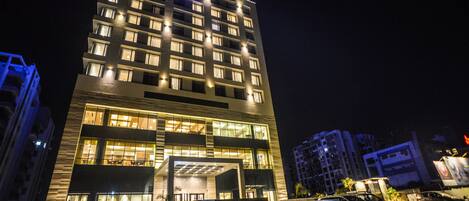 Un hotel de varios pisos con fachada moderna, amplias ventanas y un estacionamiento con varios automóviles.