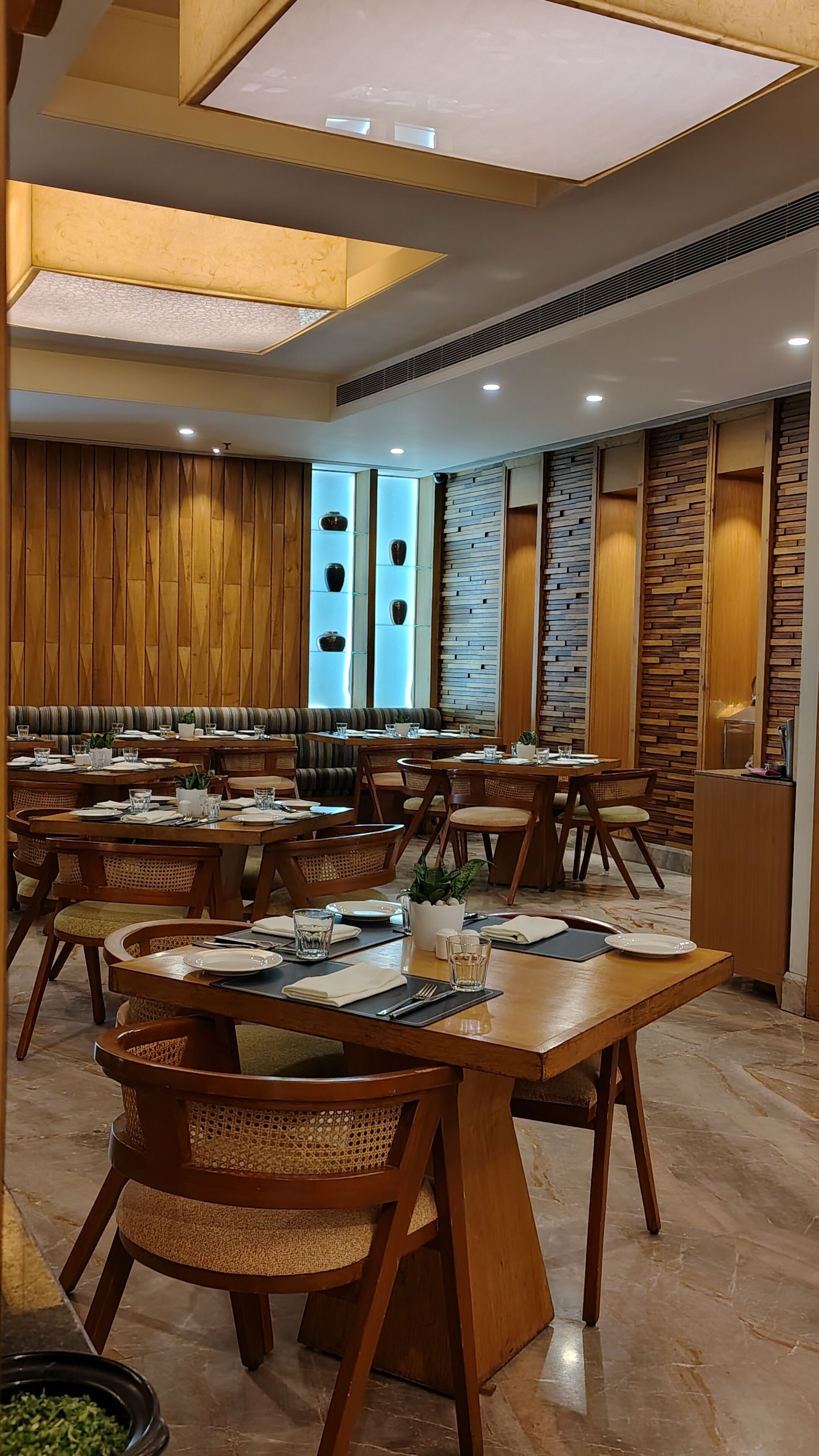 A restaurant with multiple wooden tables and chairs, a high ceiling with recessed lighting, and a stone wall with a wooden panel design.
