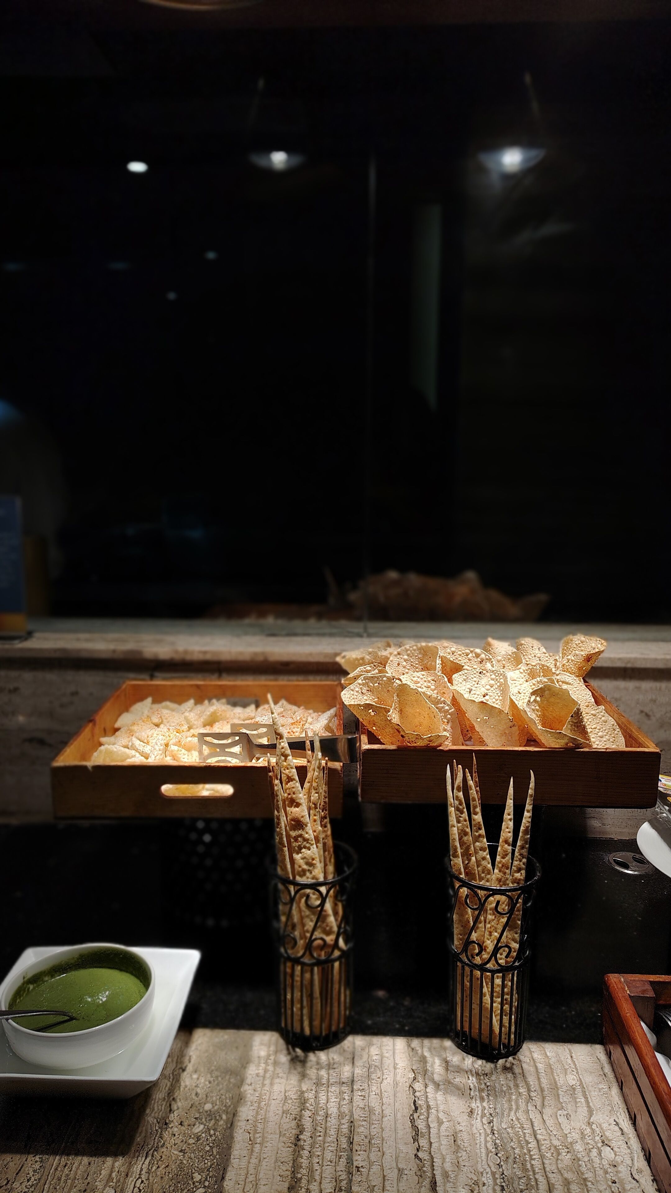 A wooden table with a variety of food items, including a bowl of green sauce and several containers of breadsticks.