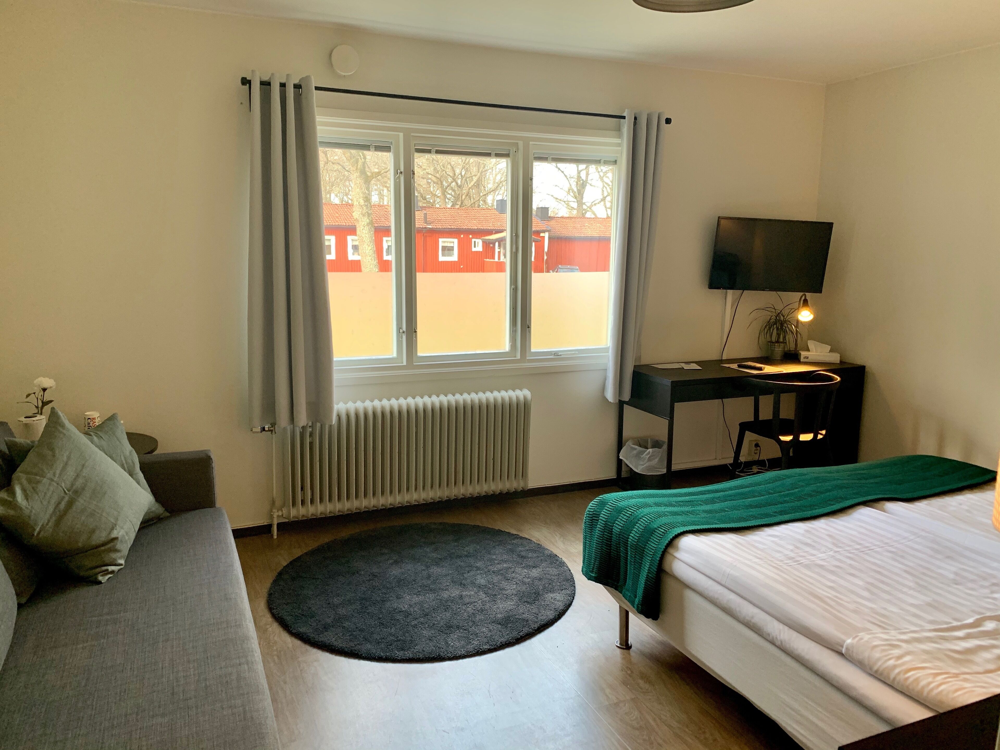 A hotel room with a bed, a sofa, a desk with a TV, a radiator, and a window with curtains.