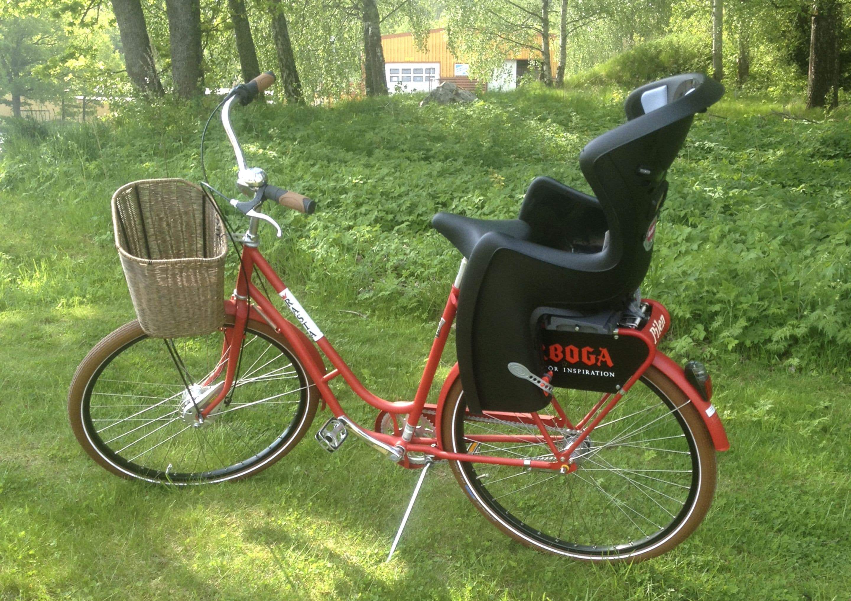 A red bicycle with a basket and a black seat, parked on grass with trees and a building in the background.
