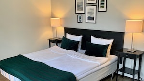 Una cama bien hecha con un plaid verde, dos mesitas de noche con lámparas y una pared con cuadros.
