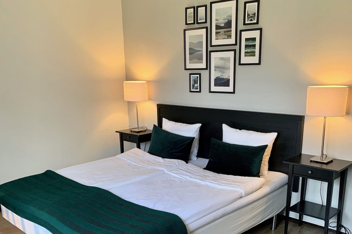 Una cama bien hecha con un plaid verde, dos mesitas de noche con lámparas y una pared con cuadros.