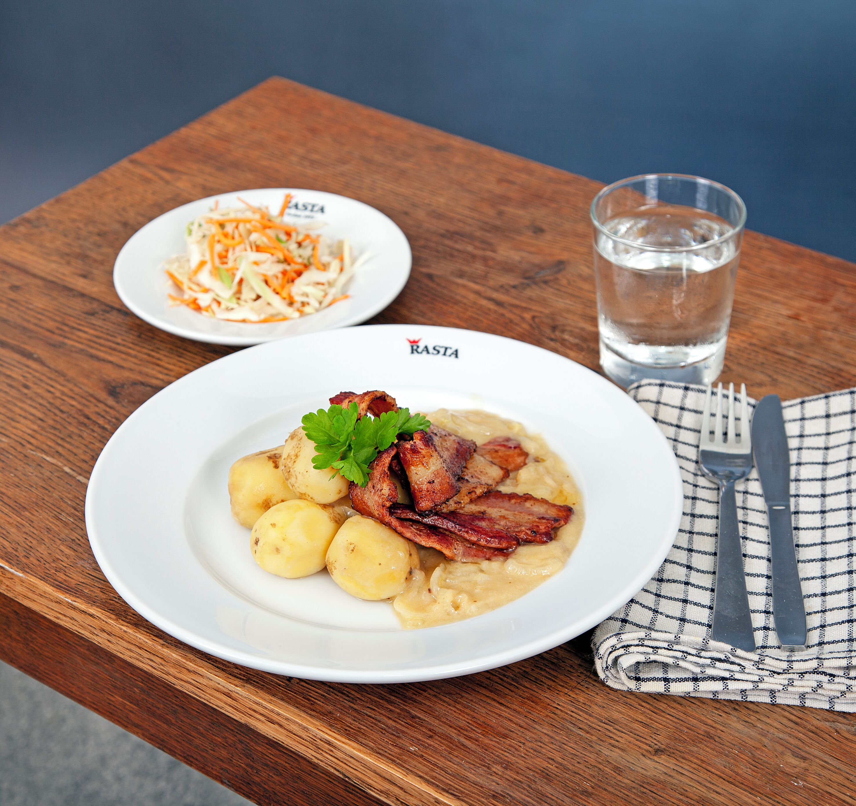 Un plato de comida con patatas y tocino, un vaso de agua y un tenedor sobre una mesa de madera.
