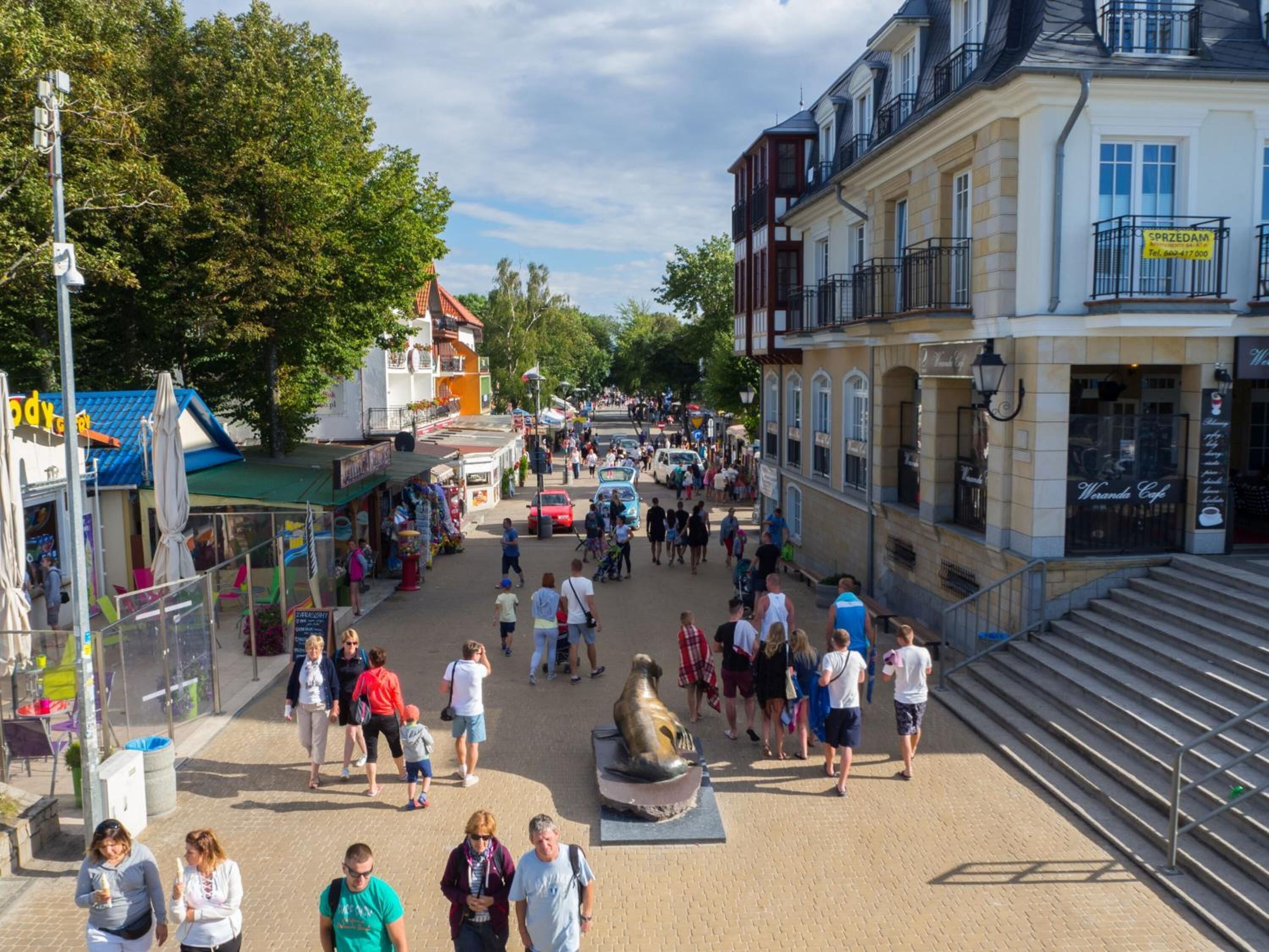 Eine belebte Fußgängerzone mit Geschäften und Cafés, einer Statue und einer Treppe, die zu einem Gebäude führt.