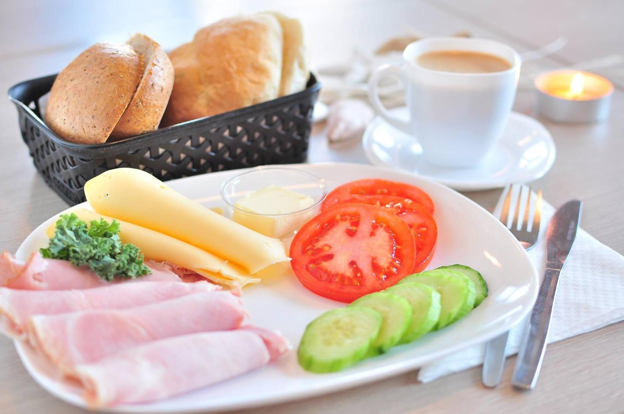 A breakfast plate with ham, cheese, tomato, cucumber, and bread, along with a cup of coffee and a candle on a table.