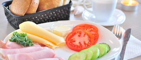 A breakfast plate with ham, cheese, tomato, cucumber, and bread, along with a cup of coffee and a candle on a table.