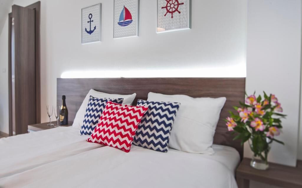 Un lit bien fait, avec une literie blanche, deux coussins décoratifs à motifs en zigzag, une bouteille de vin et un vase de fleurs posés sur une table de chevet.