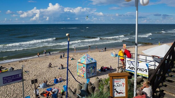 Ein Strand mit Menschen, einem Rettungsturm und einem Schild, das Sportbrillen wirbt.