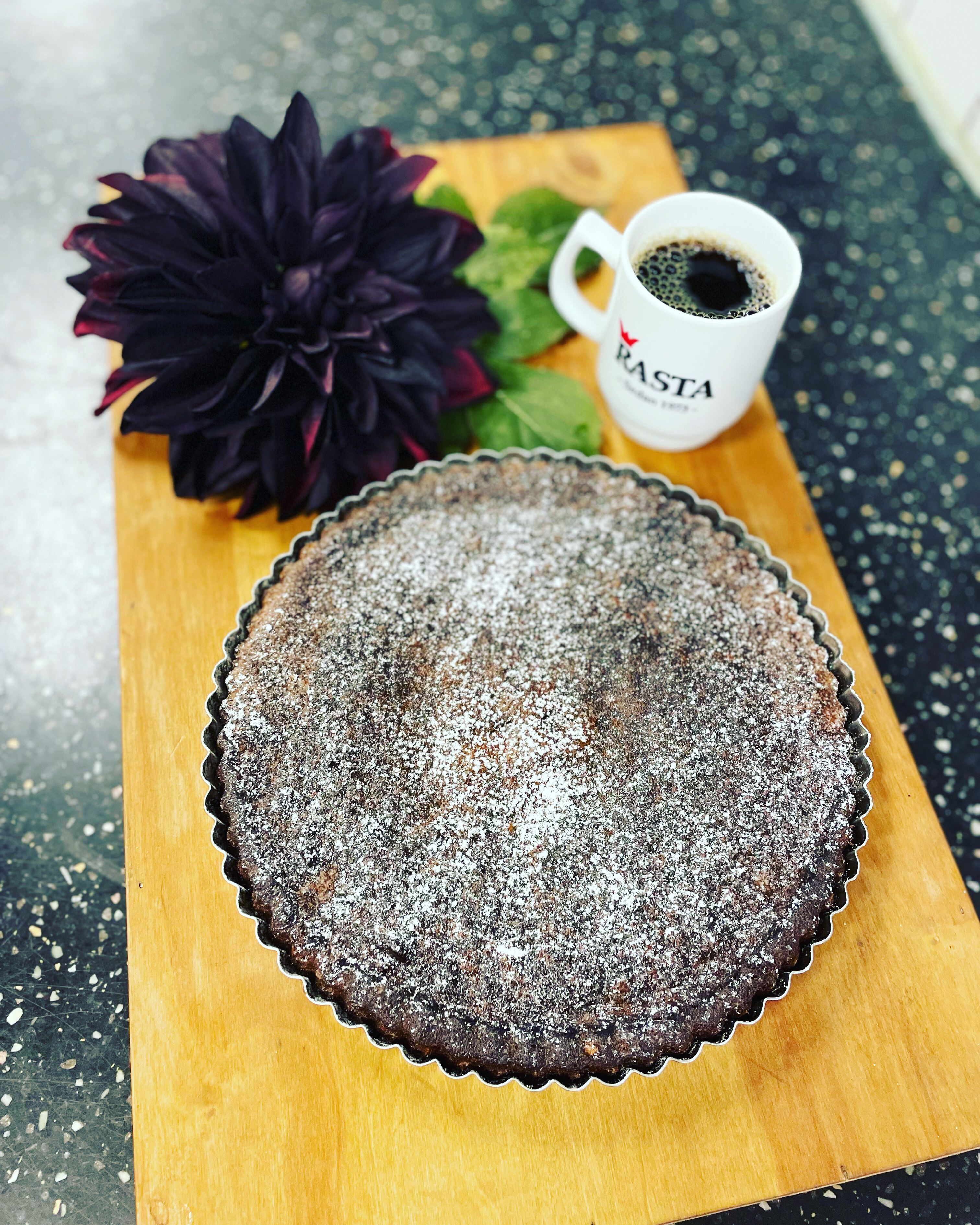 Un pastís de xocolata amb sucre glas sobre una taula de fusta, una flor de color morat fosc i una tassa de cafè amb el text 