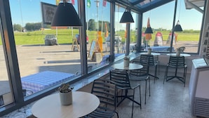 An indoor seating area with round tables and chairs, large windows offering a view of a parking lot and green fields.