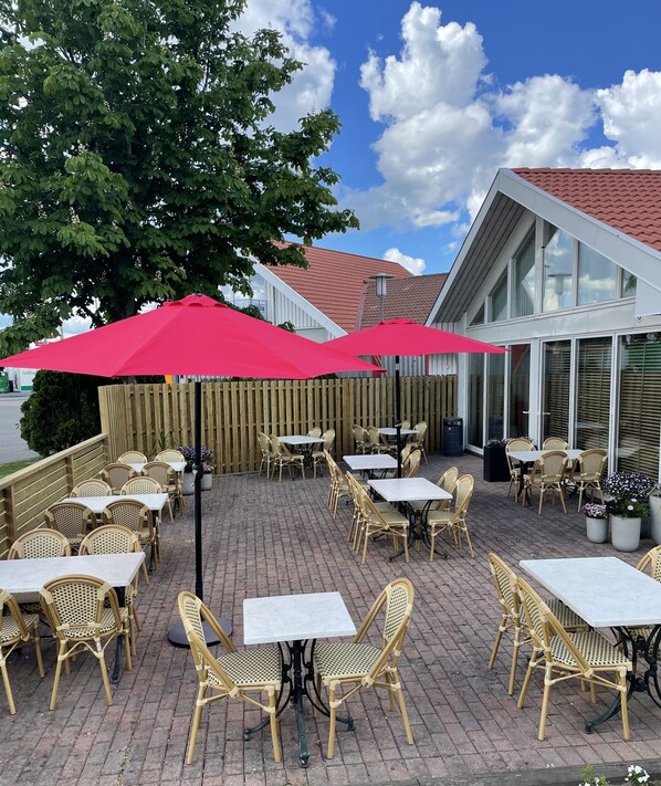 An outdoor patio with wicker tables and chairs, red umbrellas, and a brick floor.