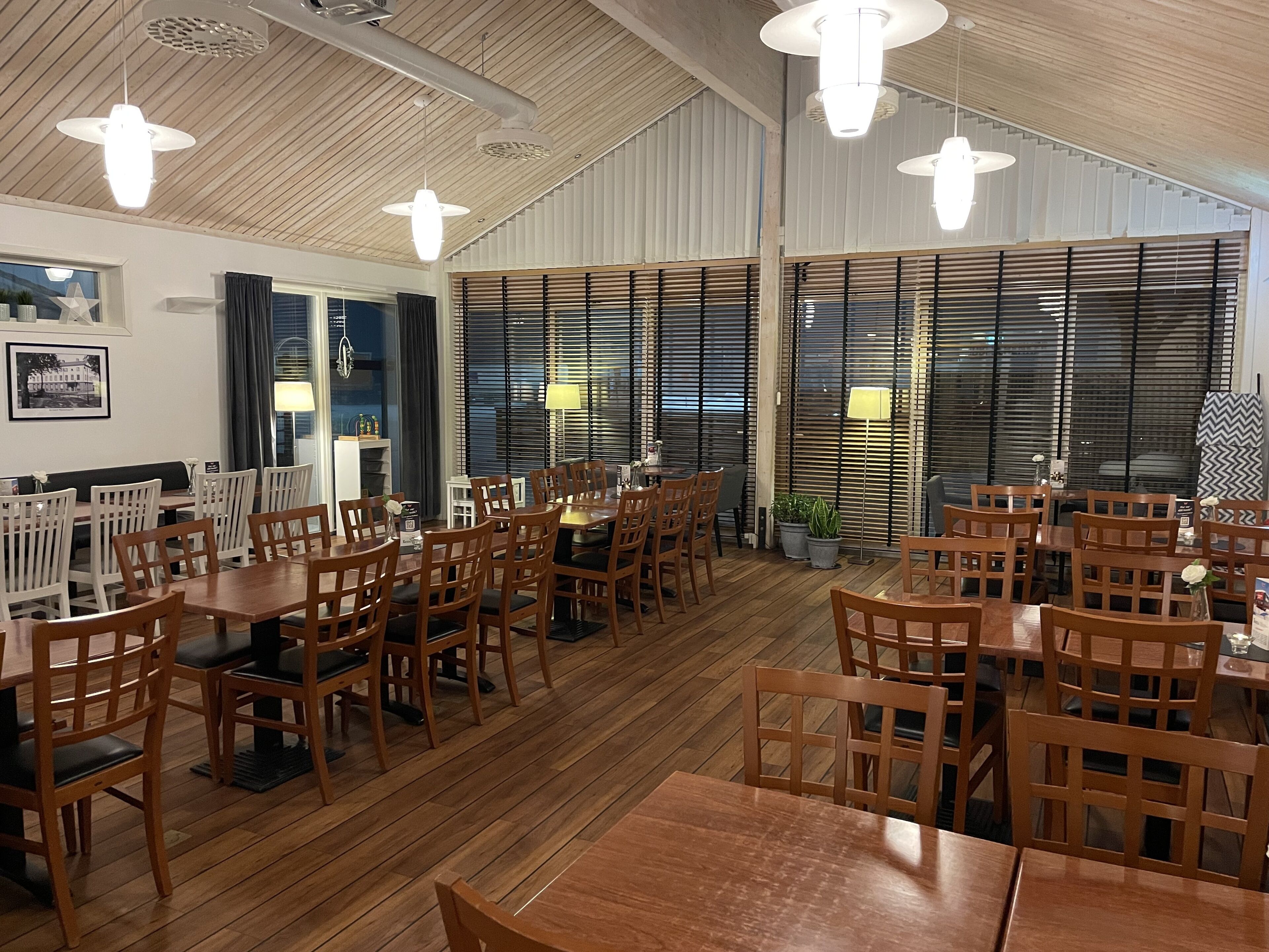 En restaurant med træborde og stole, store vinduer med persienner og hængende pendellamper.
