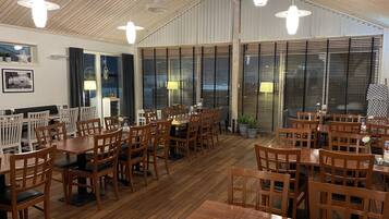En restaurant med træborde og stole, store vinduer med persienner og hængende pendellamper.