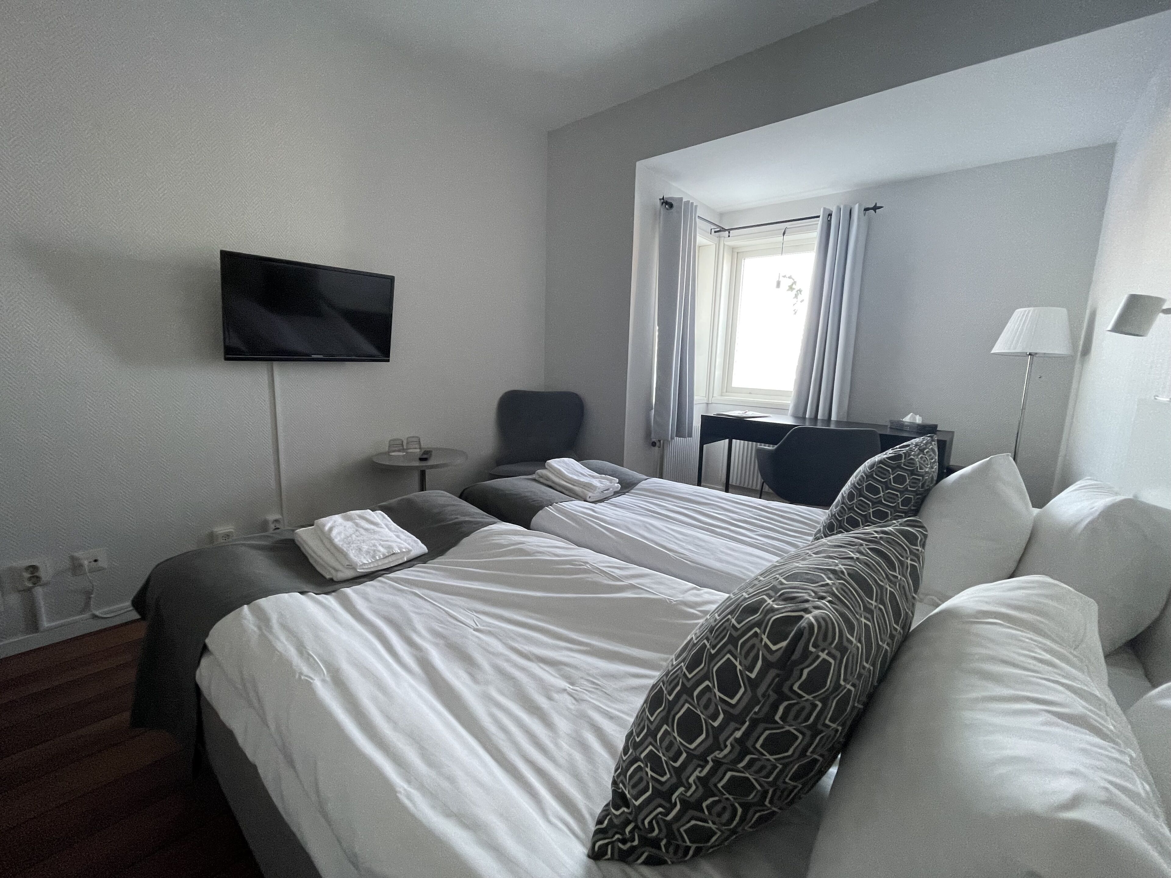 A hotel room with two beds, a flat-screen TV mounted on the wall, a desk with a chair, a lamp, and a window with curtains.