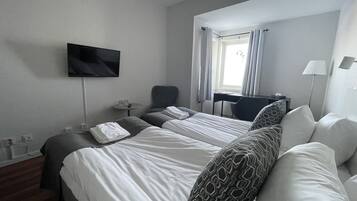 A hotel room with two beds, a flat-screen TV mounted on the wall, a desk with a chair, a lamp, and a window with curtains.