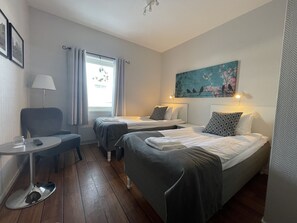 A hotel room with two single beds, a small table with a lamp, a chair, and a window with curtains.