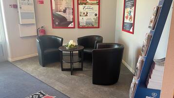 A hotel lobby with a reception desk, a seating area with two chairs and a table, and a display of brochures.