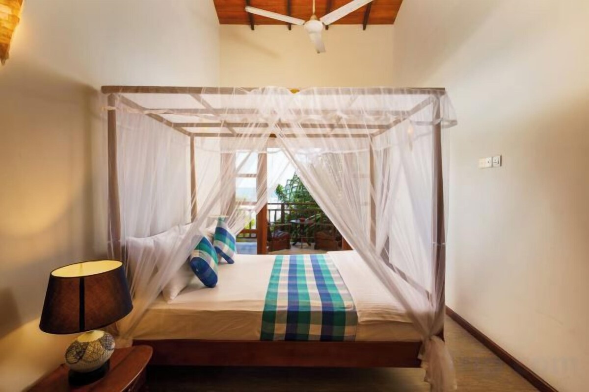 A room with a bed under a canopy, a bedside table with a lamp, and a view of the outdoors.