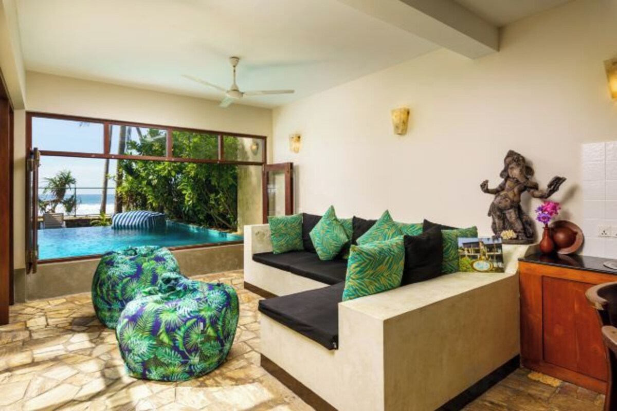 Un espace de vie moderne comprenant un canapé, des fauteuils en forme de poire, une petite table et une statue. Une grande fenêtre offre une vue sur une piscine et sur la verdure environnante.