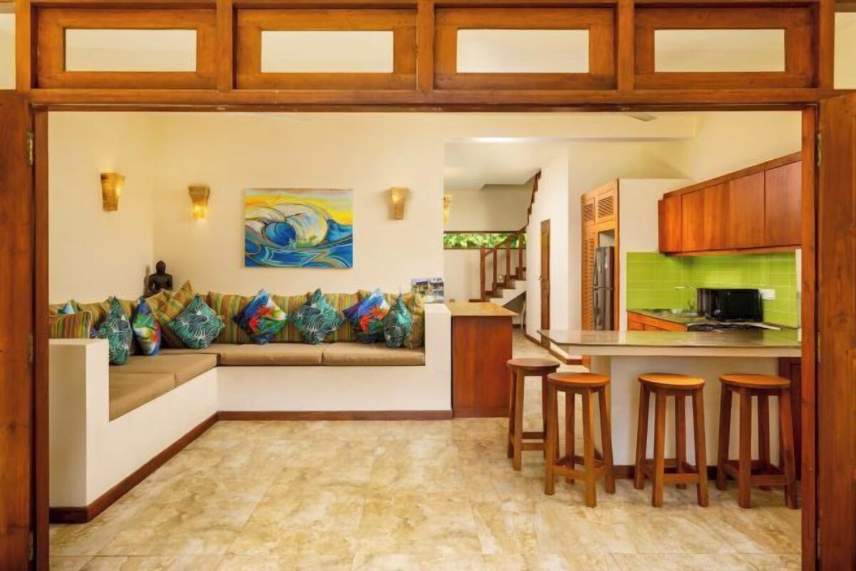 A kitchen with a wooden bar stool area, a green backsplash, and a painting on the wall.