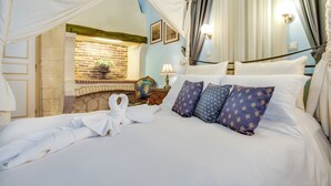 A neatly made bed with white linens and a canopy, a stone fireplace, and a nightstand with a lamp.
