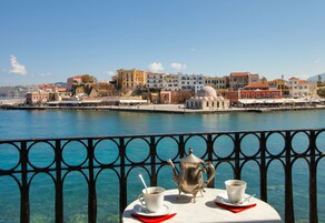 Ένα τραπέζι με μια τσαγιέρα και φλιτζάνια σε ένα μπαλκόνι με θέα στην παραλία, με κτίρια να διακρίνονται στο βάθος.