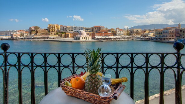 A balcony with a wicker basket containing fruits and a bottle, overlooking a waterfront with buildings and a dome structure.