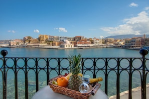 A balcony with a wicker basket containing fruits and a bottle, overlooking a waterfront with buildings and a dome structure. - Casa Leone Hotel (Chania)