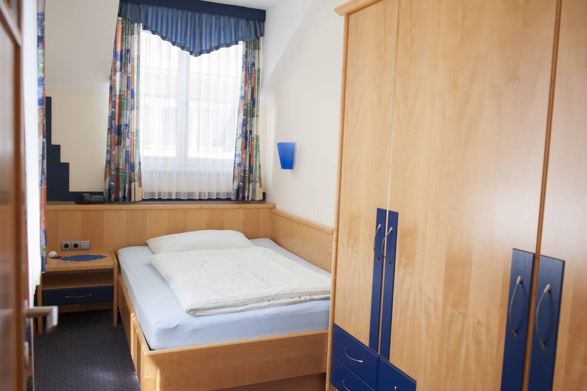 Un dormitorio con cama, armario de madera, mesita de noche, escritorio y ventana con cortinas.