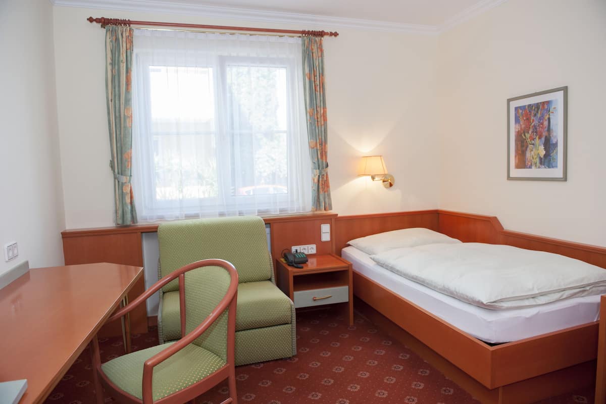 Una habitación de hotel con una cama individual, un escritorio, una silla, una mesita de noche con lámpara y una ventana con cortinas.