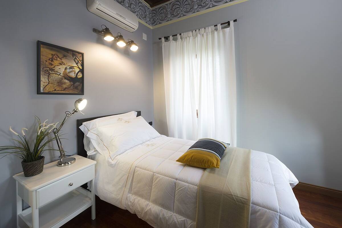 A neatly made bed with a white duvet and a yellow pillow, a bedside lamp, a small potted plant, and a framed artwork on the wall.