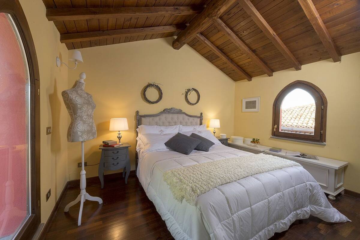 A bedroom with a wooden ceiling, a bed with white bedding, two wall sconces, a dresser, a window with a view, and a mannequin.