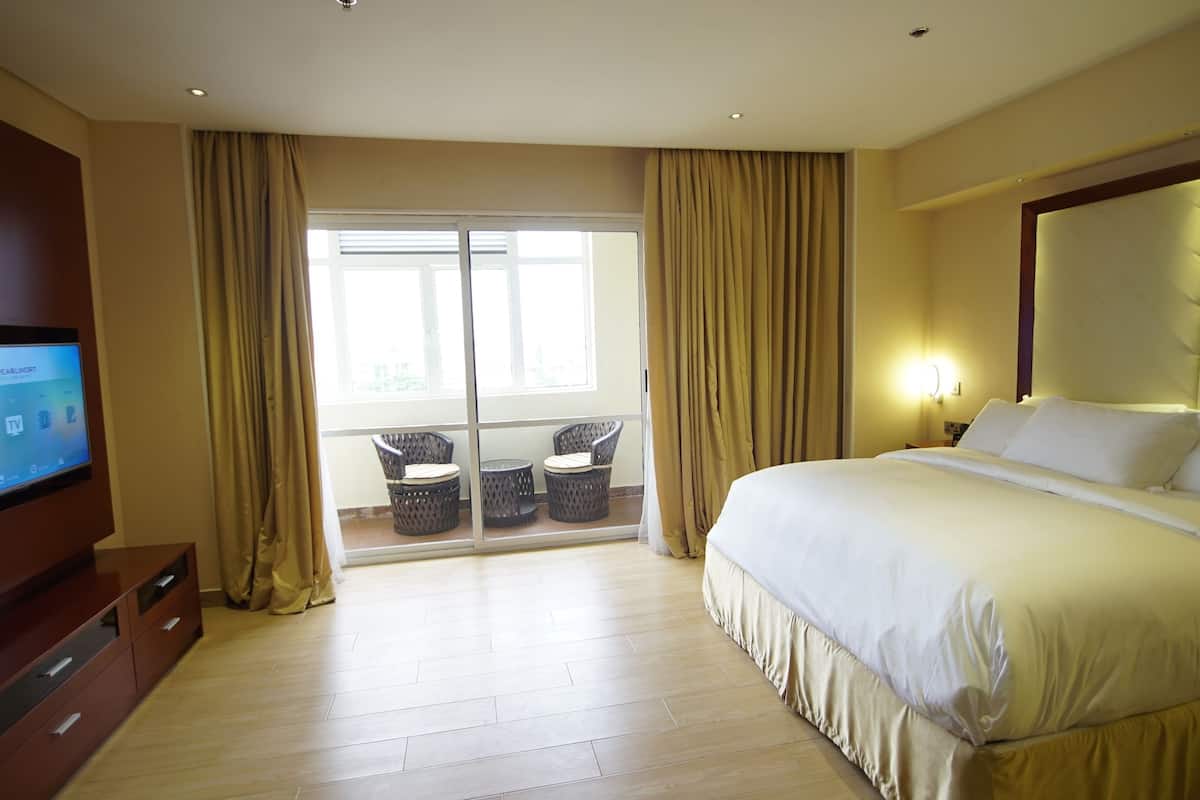 A hotel room with a large bed, a TV on a wooden cabinet, and a balcony with wicker chairs.