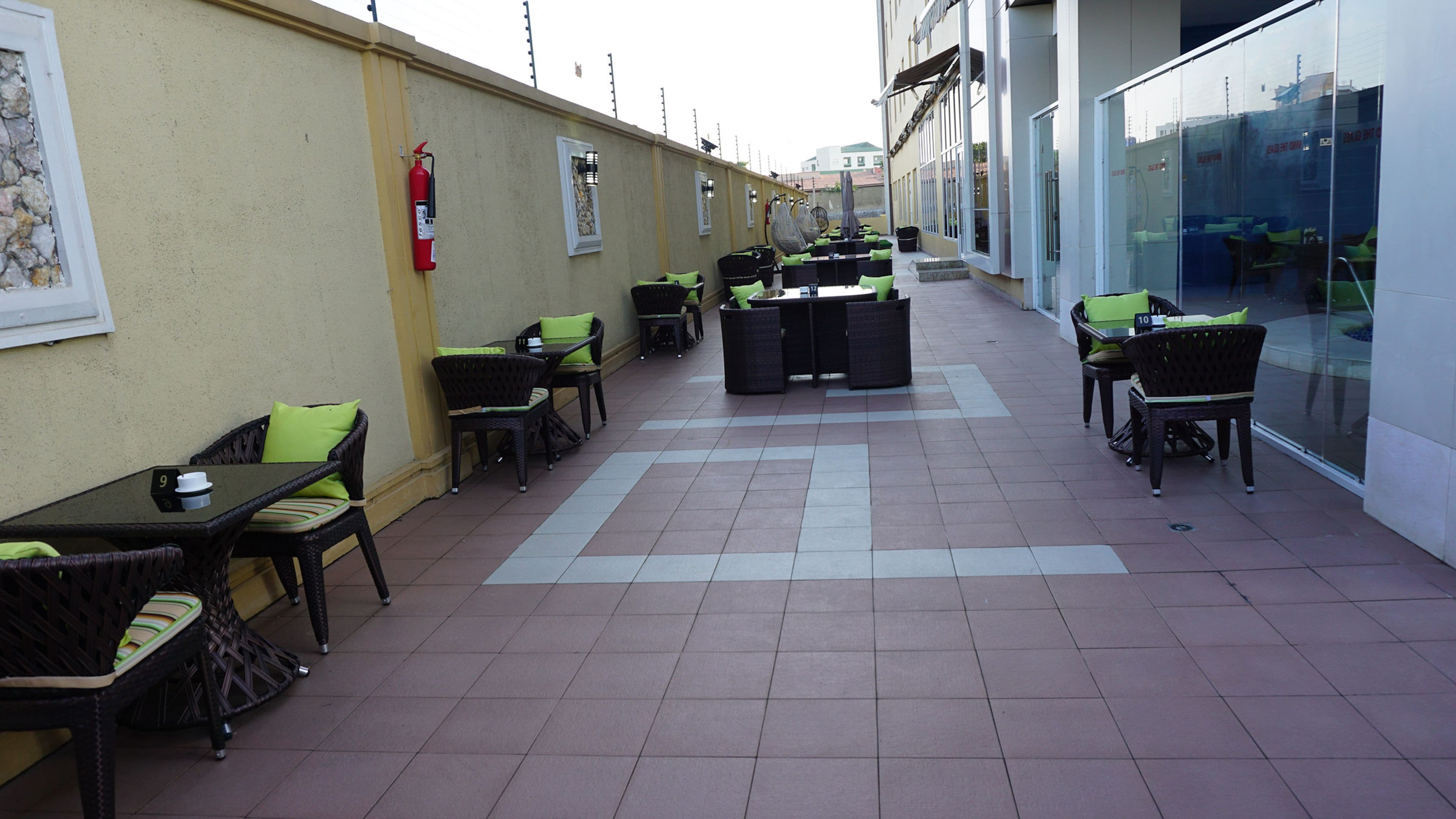 Eine Terrassengruppe mit mehreren Tischen und Stühlen, ein Feuerlöscher an der Wand und ein Glasfachhaus im Hintergrund.