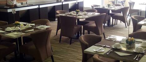 A hotel dining area with multiple tables set for dining, a kitchen area with a counter, and a view through large windows.