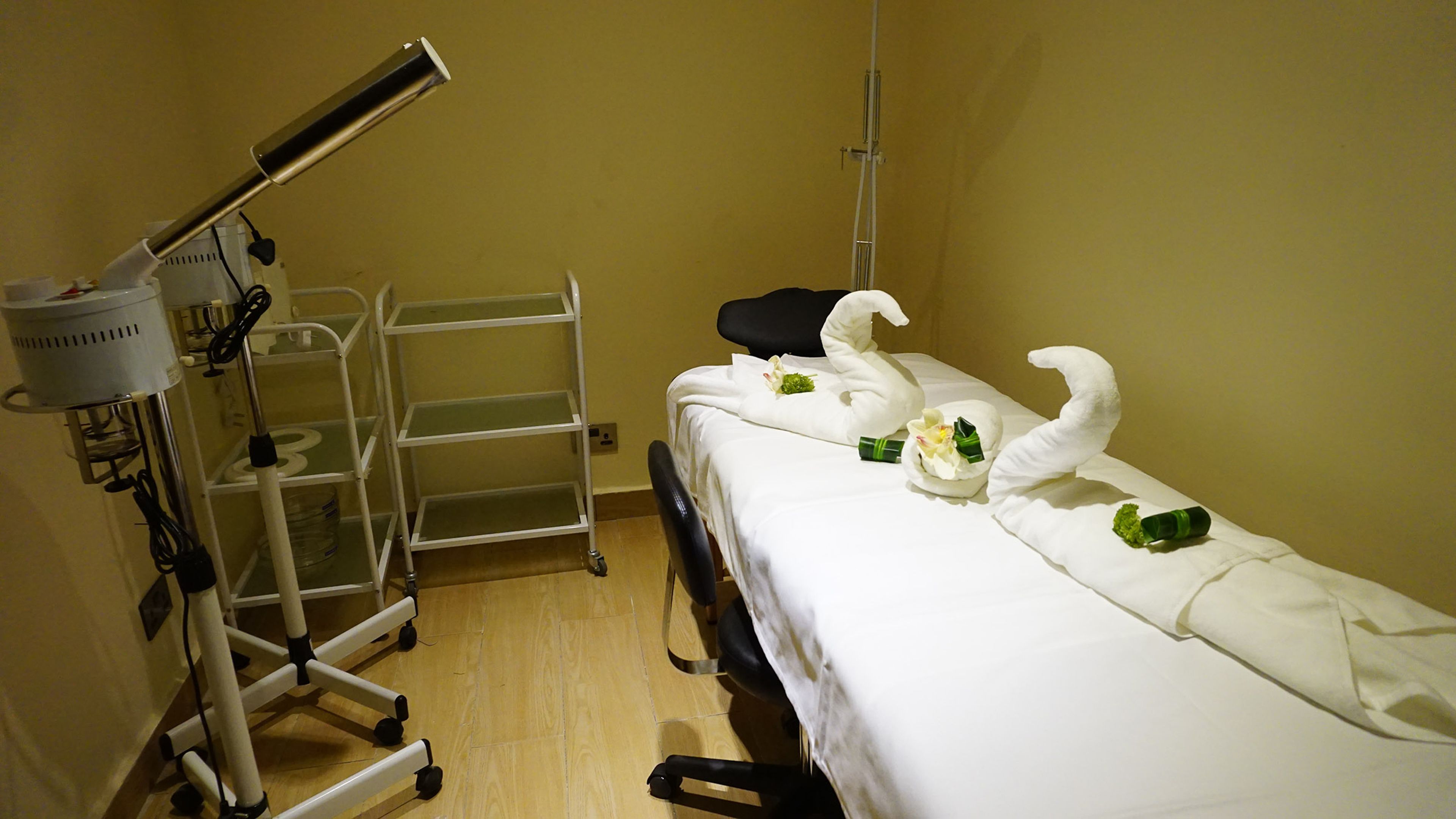 Hotelkamer met een bed, een witte handdoek met zwanen, een klein boeket bloemen en een medische lamp.