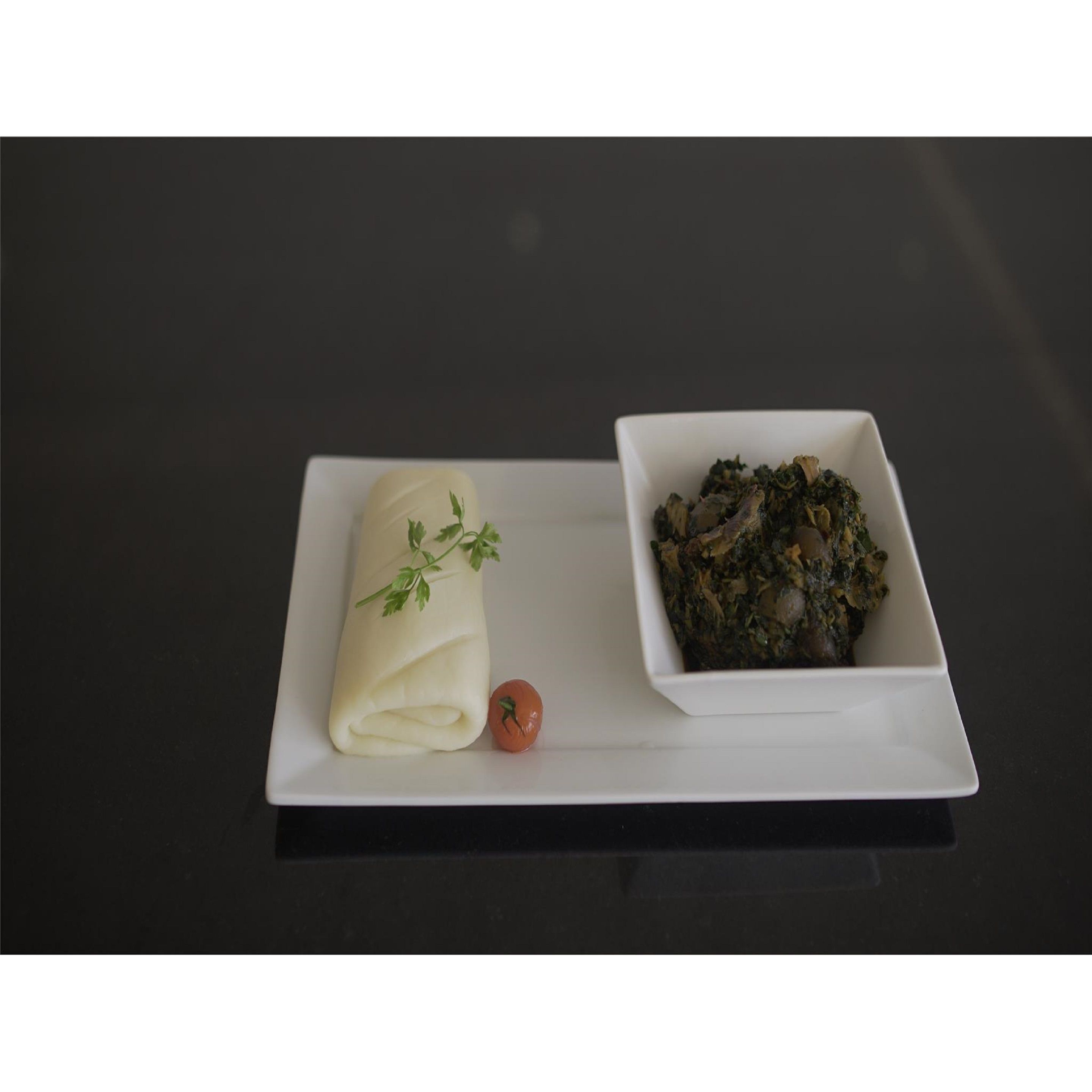 Un plato blanco con un alimento enrollado y un tazón de vegetales verdes, acompañado de un tomate pequeño sobre una superficie oscura.