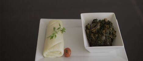 Un plato blanco con un alimento enrollado y un tazón de vegetales verdes, acompañado de un tomate pequeño sobre una superficie oscura.