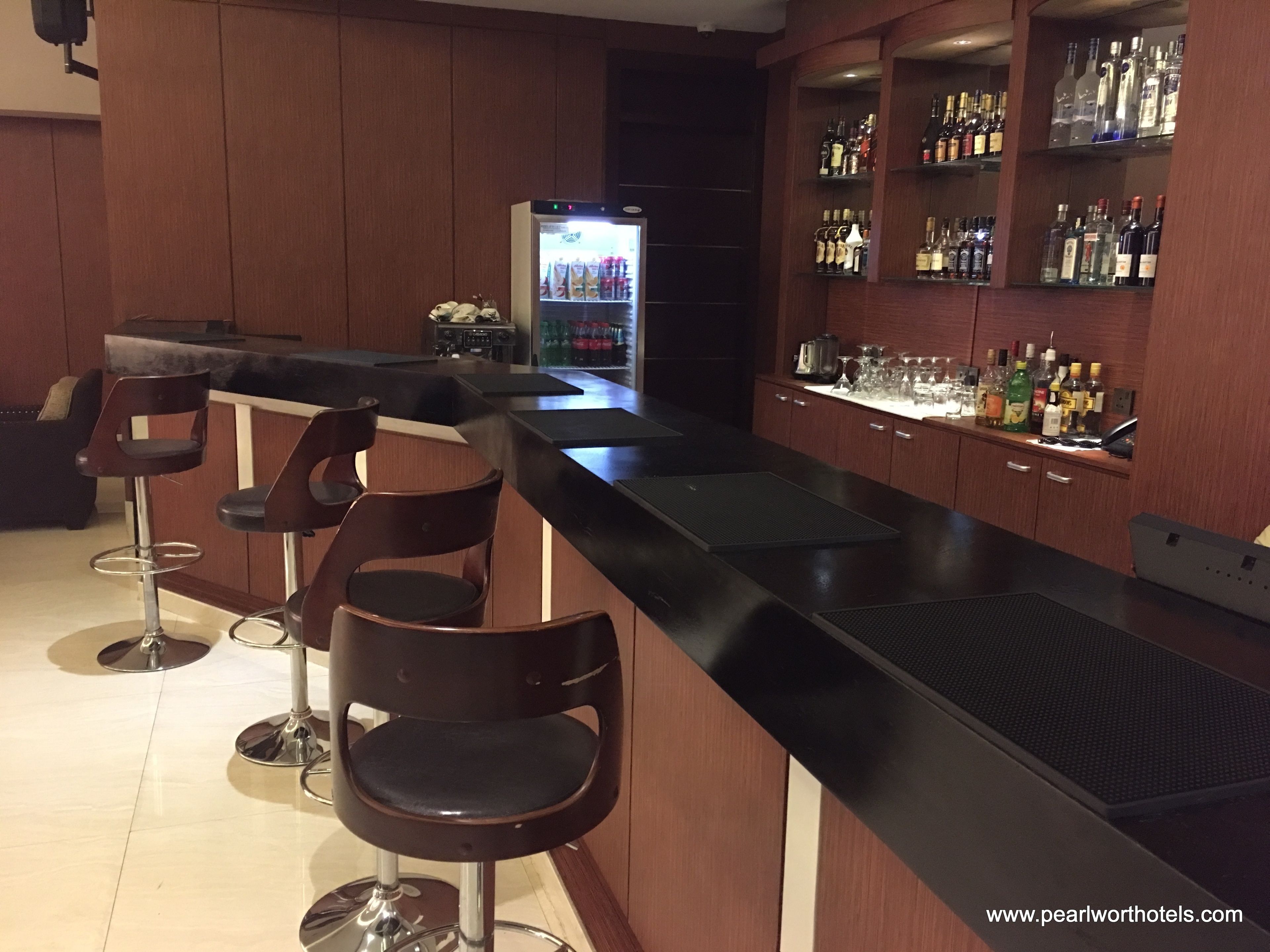 A bar area with a long counter, three stools, a refrigerator stocked with beverages, and shelves with various bottles.