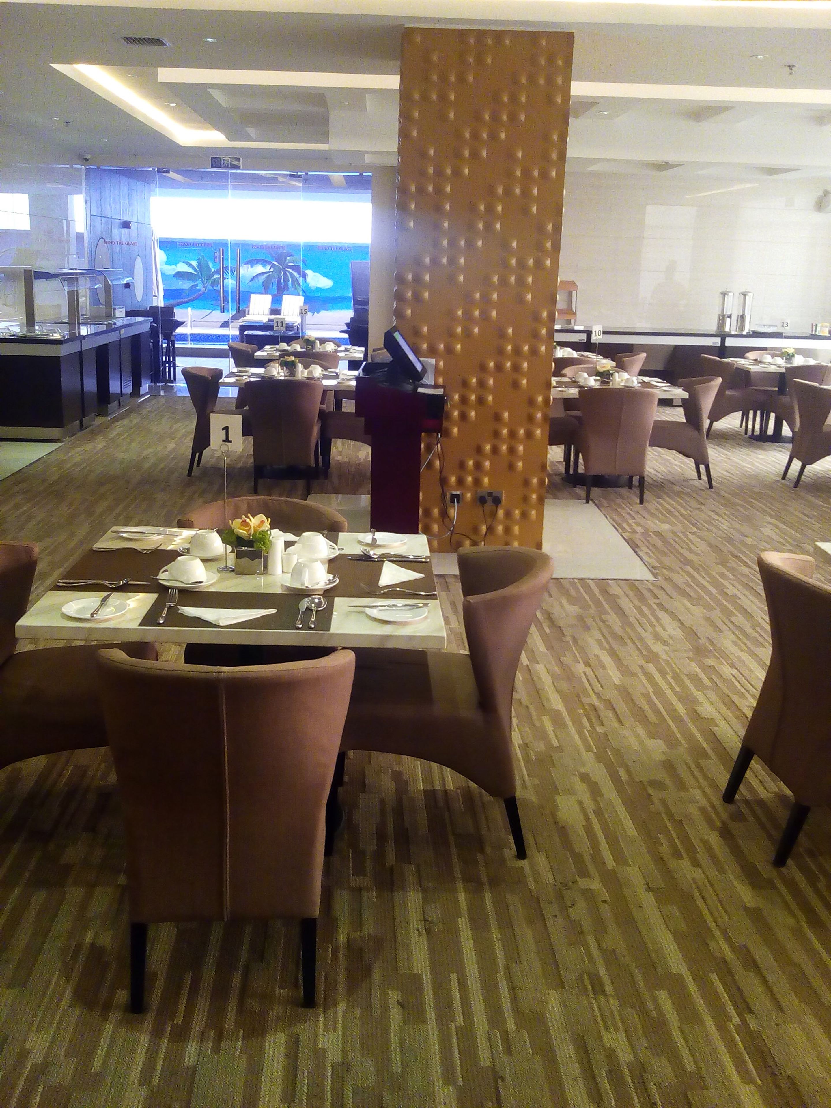 A hotel dining area with tables set for four, a bar in the background, and a large screen displaying a movie.