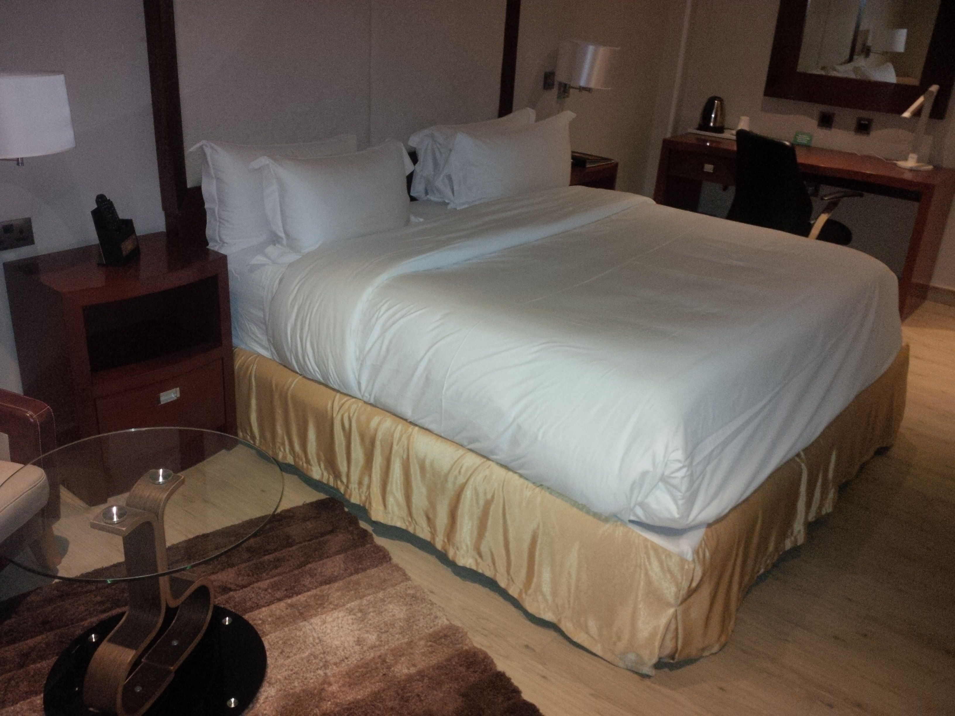 A hotel room with a large bed, a desk, a chair, a lamp, and a small table with a guitar.
