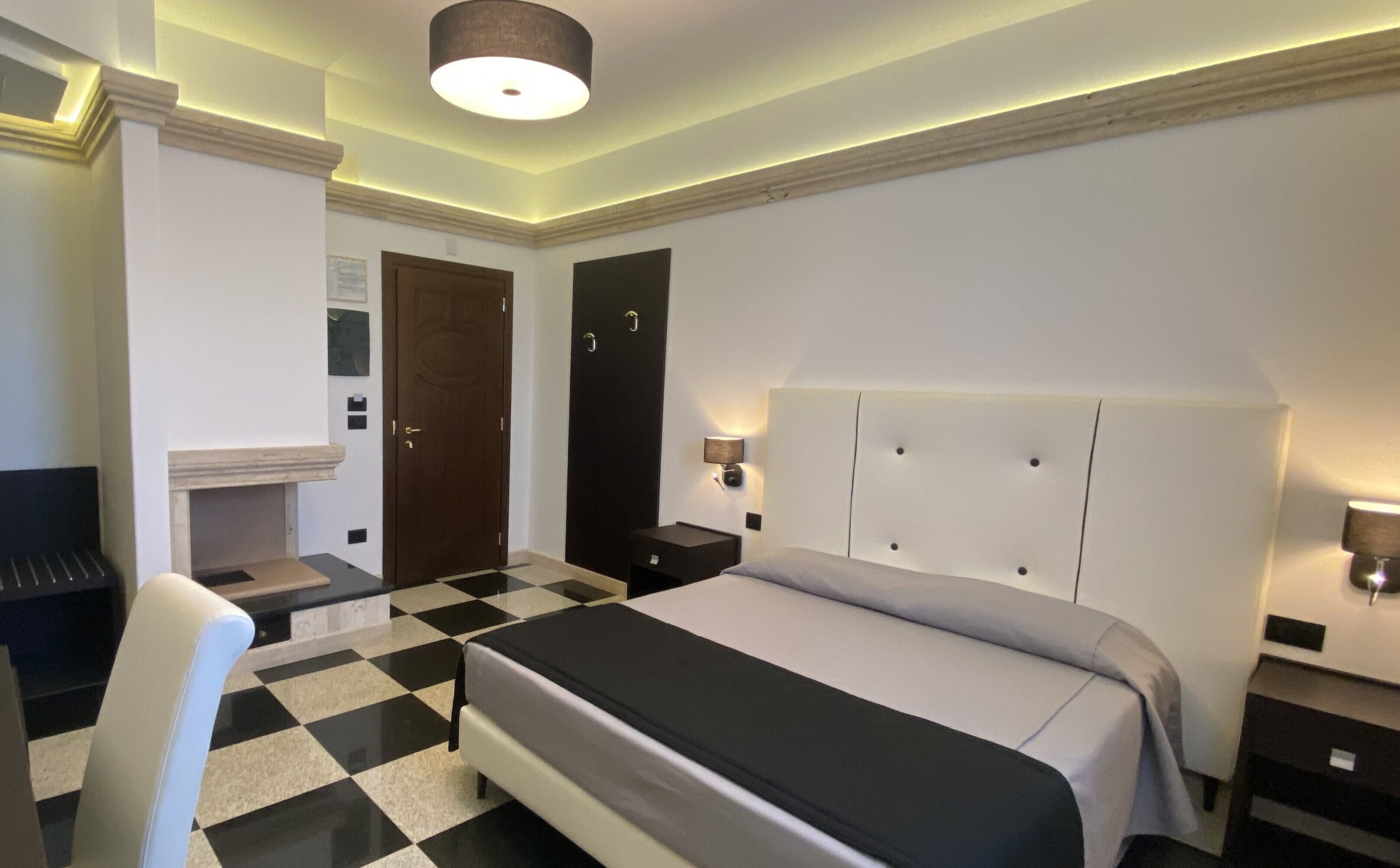 A hotel room with a checkered floor, a bed with a headboard, a desk, a chair, a door, and a wall-mounted lamp.