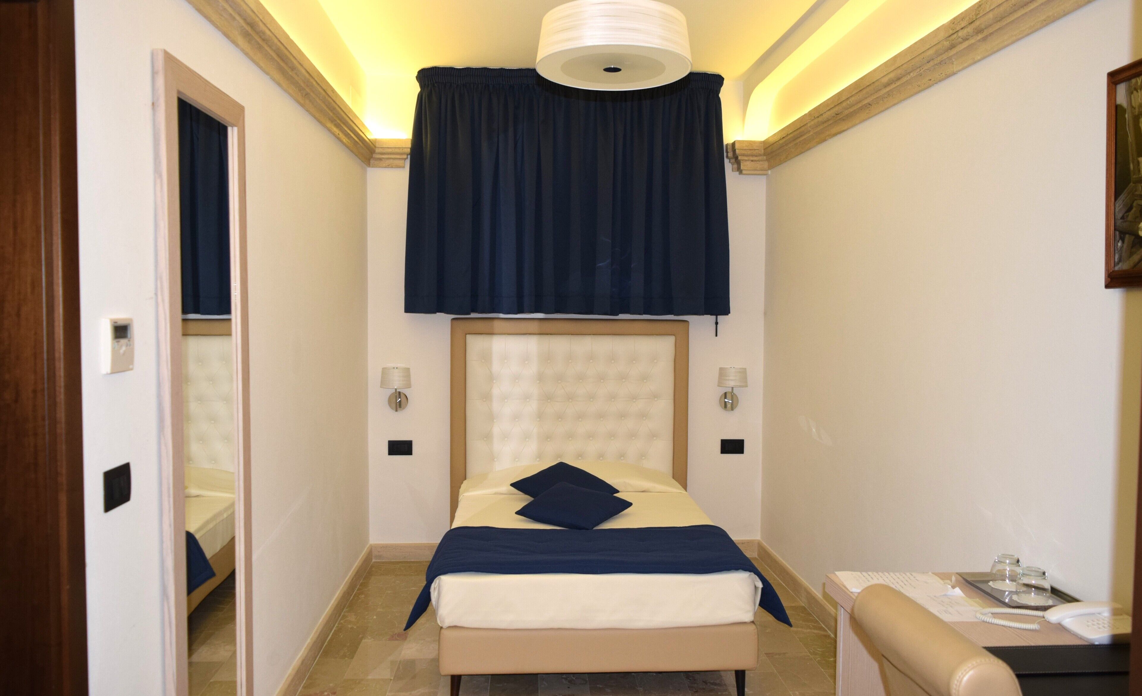 Una camera d'albergo modesta con un letto, abat-jour, un piccolo tavolino con un telefono e uno specchio.