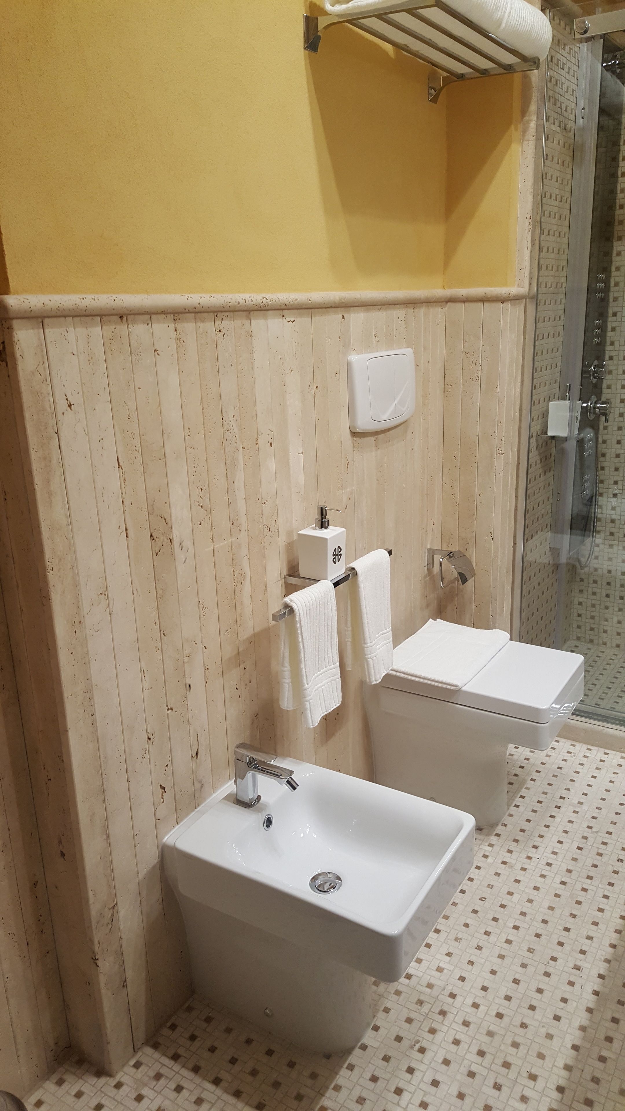 Une salle de bain avec un lavabo blanc, des toilettes et une douche. On y trouve un porte-serviettes avec des serviettes et un distributeur de savon fixé au mur.